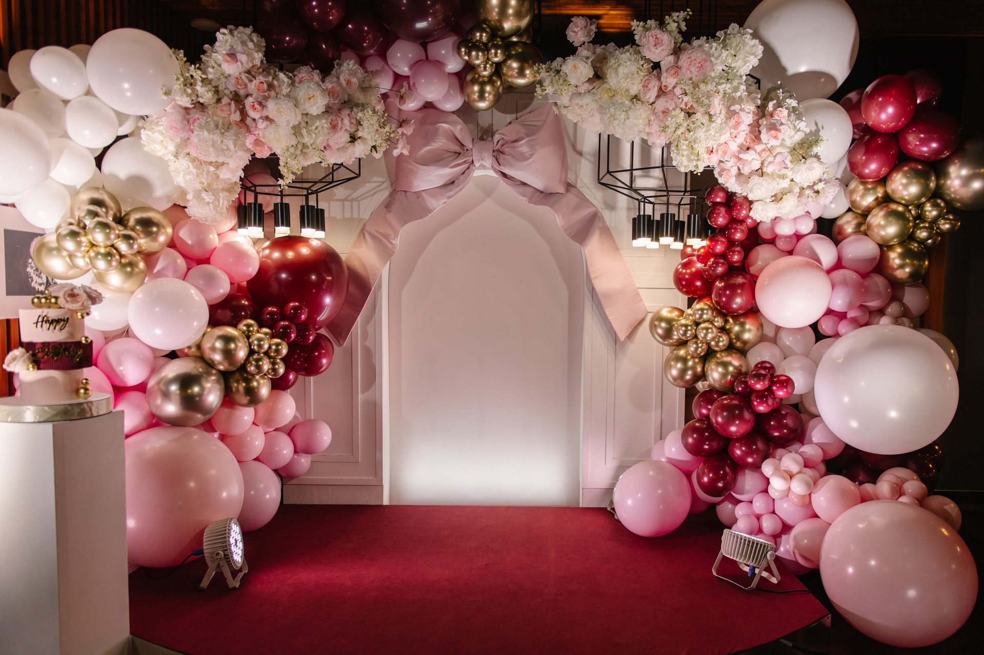 Birthday party decorations. Three-tiered cake with pink roses, Happy Birthday text topper, and gold decor.  Trendy burgundy Cake on a background of balloons on arch. Celebration concept.