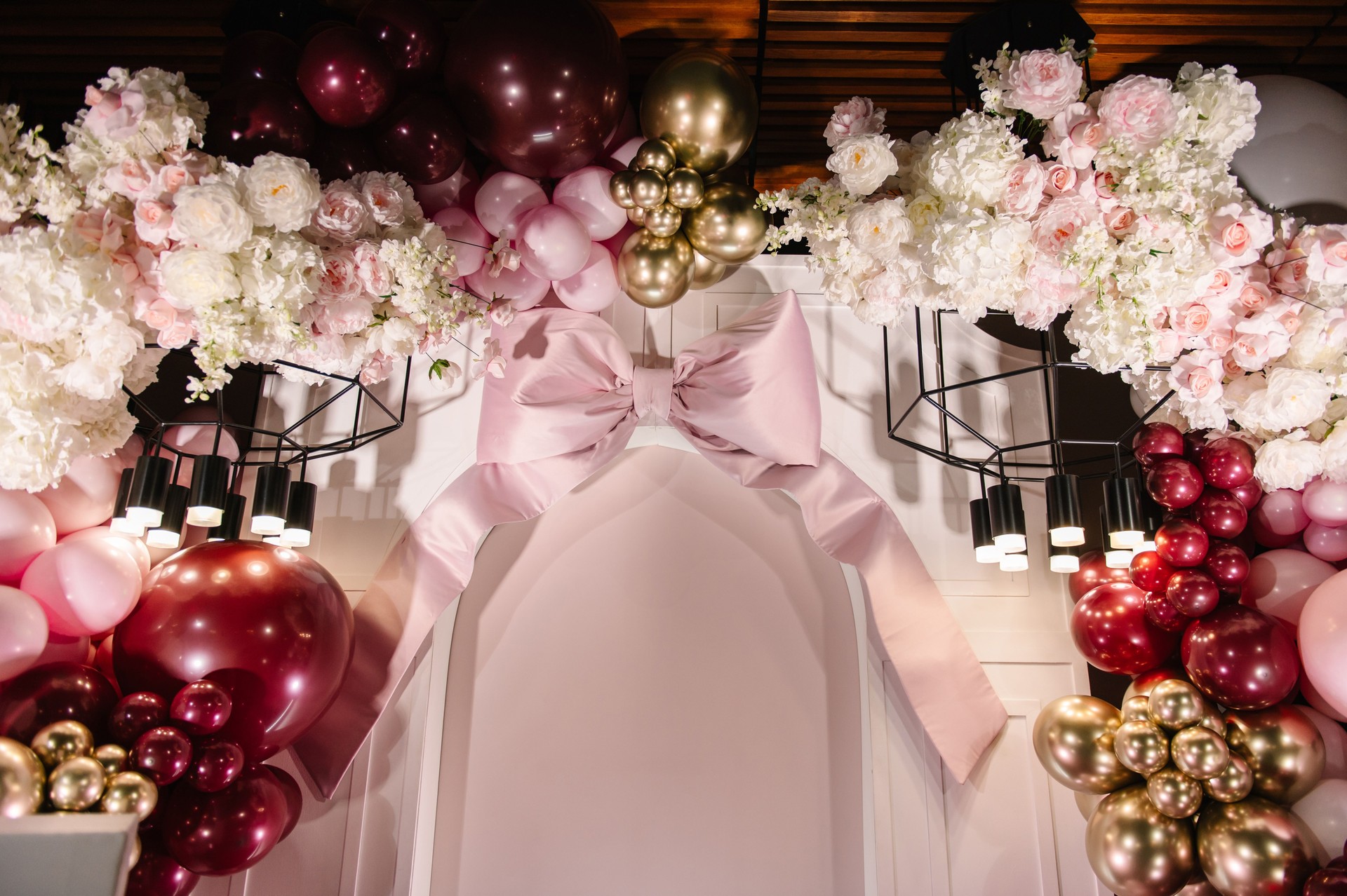 Big pink bow. Arch was decorated with pink, gold, burgundy balloons, flowers and a big bow. Photo wall decoration space or place for text. Reception at birthday party. Luxury decor for woman parties.